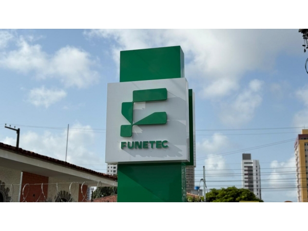 A foto mostra a chefe de gabinete, Rejane Negreiros, sentada em uma mesa de escritório, sorrindo de forma acolhedora para a câmera. Ela veste uma blusa de tom claro, com mangas longas, e está com o cabelo preso, transmitindo uma aparência profissiona